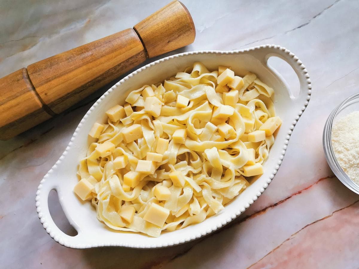 Cubes of Fontina cheese on top of cooked tagliatelle in white oval oven dish.