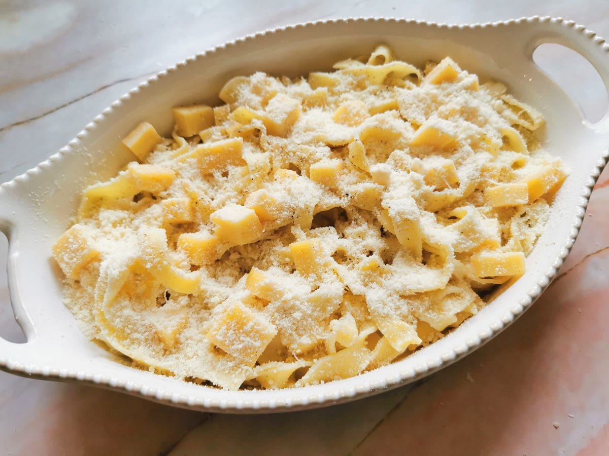 Cooked tagliatelle and Fontina cubes in white oval oven dish with Parmigiano sprinkled on top.