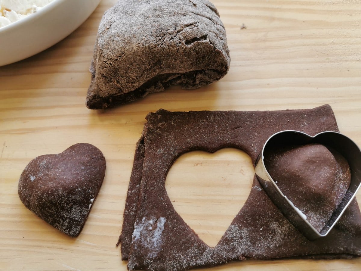 A heart shaped chocolate ravioli next to a piece of chocolate pasta dough with cookie cutter on top.
