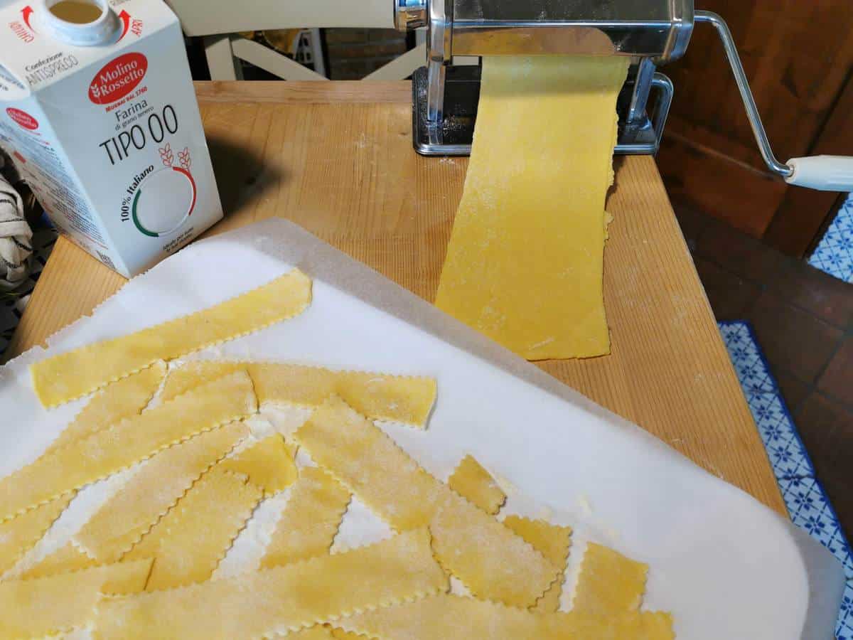 Pasta dough sheet in pasta machine and cut pasta ribbons on white tray.