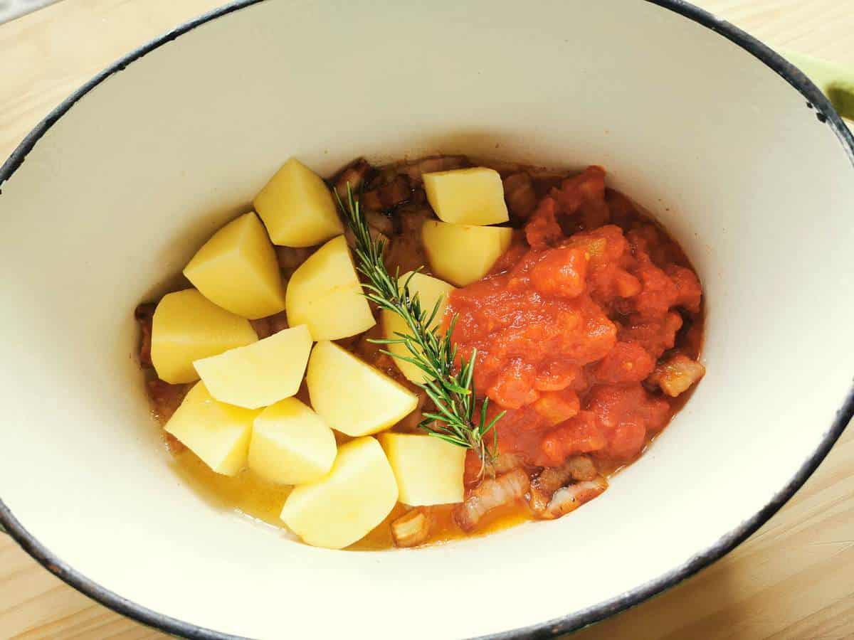 Cooked pancetta with peeled and quartered potatoes, tomato passata and rosemary in Dutch oven.