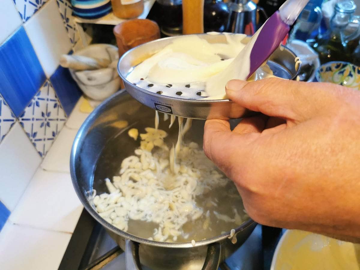 Chnèfflènè batter being pushed through a grater and falling into a pot of boiling salted water.