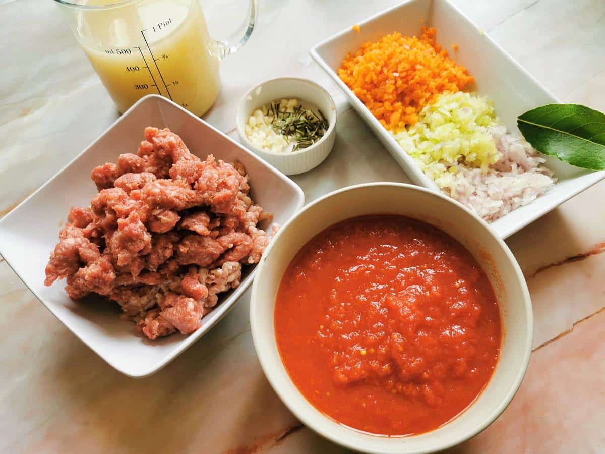 Prepared ingredients for beef sausage ragu. Ground veal and crumbled sausage meat in white bowl. Tomato passata in white bowl. Chopped vegetables and herbs in white bowls.