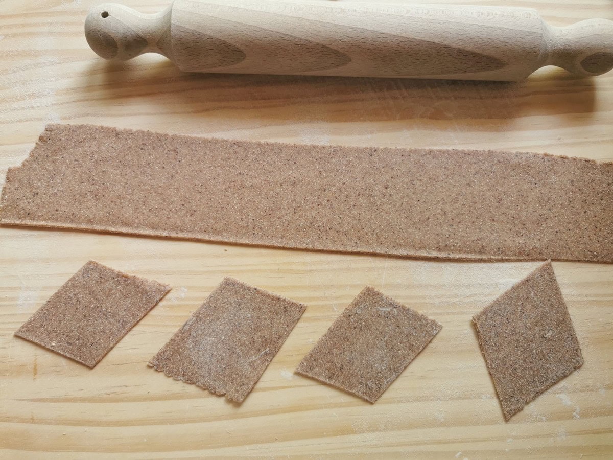 A sheet of chestnut pasta dough on wood work surface with some rhomboid shaped pieces.