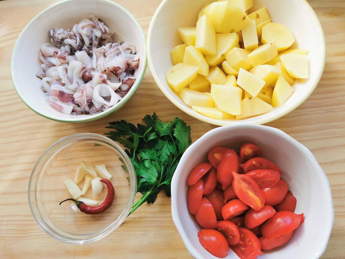 Prepared calamari, garlic, chili potatoes and cherry tomatoes in bowls. Flat parsley on a wooden board.